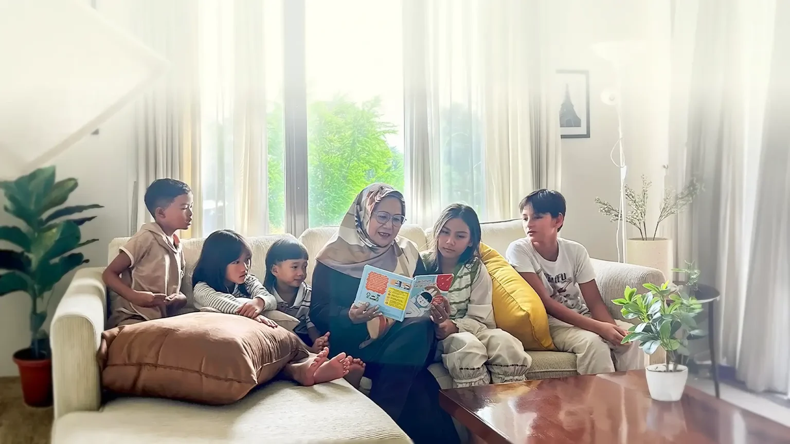 A woman reads a colorful book to a group of children seated on a sofa.
