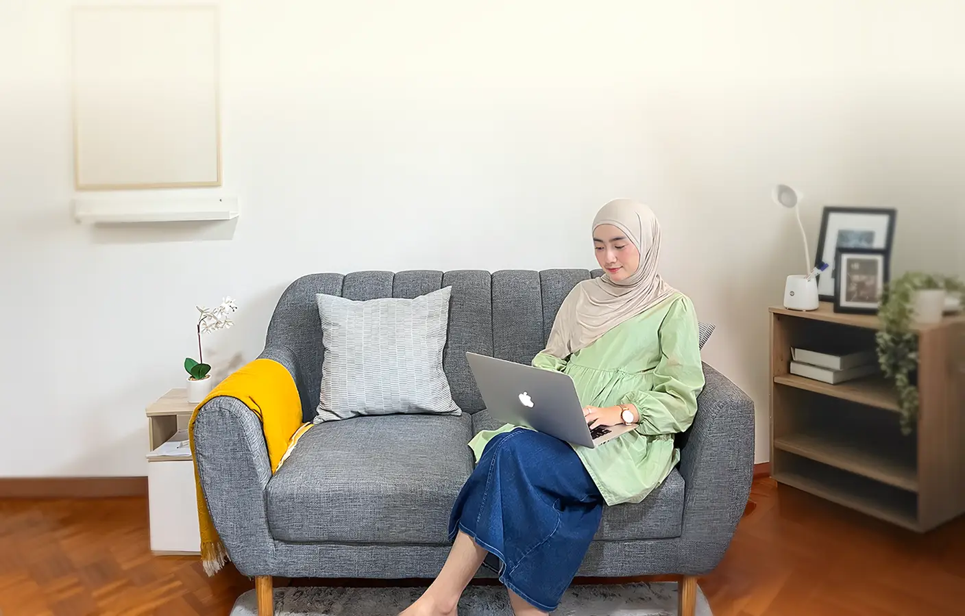 A woman in a green tunic and jeans works on a laptop while sitting on a gray sofa.